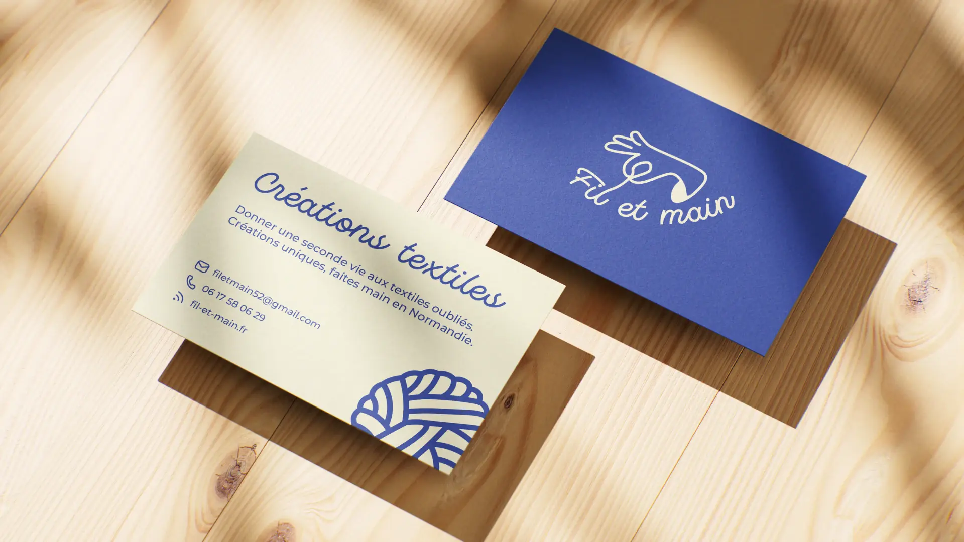 Deux cartes de visite Fil et Main sur un plan de travail en bois clair, l’une bleue au logotype, l’autre crème listant les coordonnées.